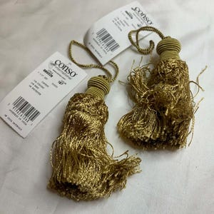 May include: Two gold decorative tassels with matching cord loops and tags. The tassels are made of a shiny, fringed material. The tags are white with black text and a barcode.