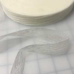 May include: A roll of off-white ribbon sits above a length of the same ribbon, unrolled and curved. The ribbon has a slightly textured appearance, and the roll is tightly wound. The background is a white surface.