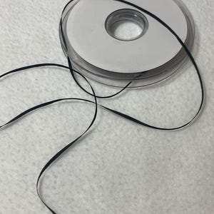 May include: A spool of black ribbon with a white center. The ribbon is unwound and lying on a white surface.