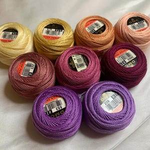 May include: A collection of nine spools of DMC cotton thread in various shades of yellow, pink, and purple. Each spool has a label with a product code. The threads are arranged on a white surface.