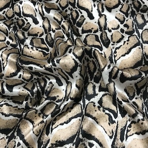 May include: A close-up of a fabric with a snakeskin pattern. The fabric has a glossy sheen and features a repeating design of black, tan, and white shapes, creating a reptile-like texture. The fabric is draped, showing folds and shadows.