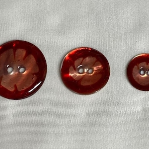 Puede incluir: Tres botones de plástico rojo con un diseño de flor y dos agujeros blancos. Los botones son de diferentes tamaños.