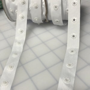 May include: White elastic with white plastic buttons. The elastic is rolled up and the buttons are evenly spaced.