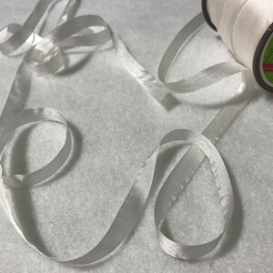 Könnte beinhalten: Eine Spule weißen Satinbandes mit einem weißen Etikett mit der Aufschrift "MADE IN JAPAN". Das Band liegt auf einer weißen Oberfläche.