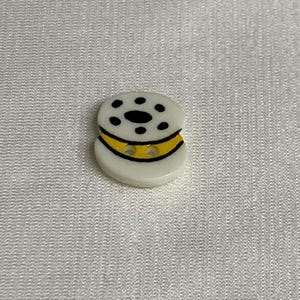 May include: White button with a black and yellow design. The button has a ring of black dots around the center and a yellow band with two holes.