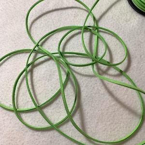 May include: Bright green leather cord, coiled and looped, against a soft, off-white background. The cord is thin and flexible, with a slightly textured surface. A black spool of the same cord is visible in the upper right corner.