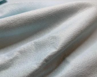 Può includere: Un tessuto in feltro azzurro con una consistenza morbida, soffice e opaca. Il materiale è drappeggiato in pieghe delicate, creando ombre e riflessi sottili sulla superficie, mettendo in risalto le fibre dense e non tessute del tessuto.