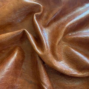 May include: A piece of medium brown leather with a natural grain texture and a glossy finish. The material is draped to create deep folds and shadows, highlighting the smooth surface and the variations in the rich, warm tan color throughout.