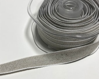 May include: A spool of natural linen ribbon with a white edge. The ribbon is wound around a clear plastic spool.