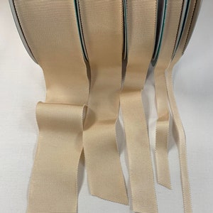 May include: Five spools of beige grosgrain ribbon, each with a different width. The ribbon is folded and fanned out, showing the different widths.