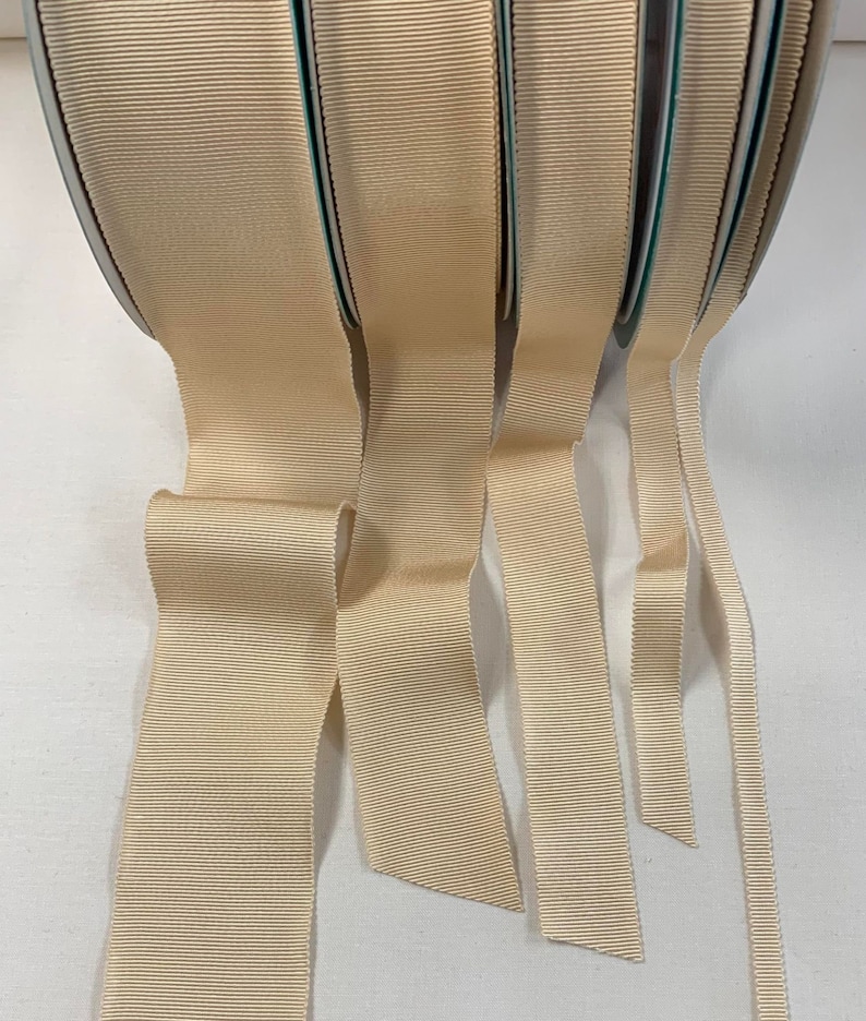 May include: Five spools of beige grosgrain ribbon, each with a different width. The ribbon is folded and fanned out, showing the different widths.
