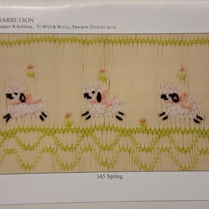 Op de afbeelding: Een close-up van een witte stof met een groen en geel geborduurd ontwerp van drie lammetjes met roze strikken. De tekst "345 Spring" is onder het ontwerp gedrukt.