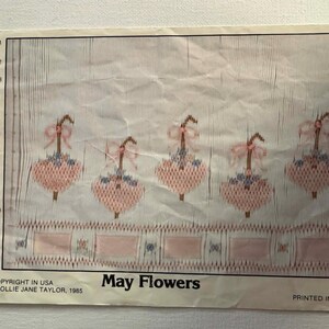 Könnte beinhalten: Ein Vintage-Stickmuster mit dem Titel "May Flowers" mit fünf rosa schirmförmigen Blumendesigns mit Schleifen und blauen Akzenten. Das Muster ist auf einem cremefarbenen Hintergrund mit einem Holzplanken-Design gedruckt. Copyright 1985.