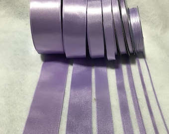 Puede incluir: Un conjunto de carretes de cinta de satén lavanda en varios anchos. La cinta está dispuesta en una pila con los extremos de la cinta que sobresalen.
