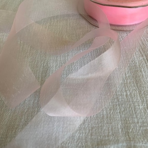 May include: A spool of pale pink sheer ribbon with a satin finish. The ribbon is wrapped around a spool with a white label that says "GREENWICH ST. 06830 TEL:(203)661-3732".