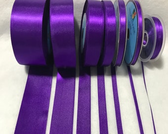 May include: A collection of purple satin ribbon spools in various widths. The ribbon is arranged in a row with the spools at one end and the ribbon extending out.