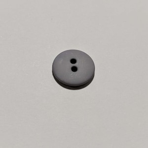 May include: A circular, grey button featuring two holes. The button is made from a smooth material and is suitable for sewing. The button's neutral colour makes it versatile for various clothing items.