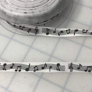 May include: White ribbon featuring a repeating pattern of black musical notes and symbols. The ribbon is spooled on a clear plastic reel. The musical notes include treble clefs, quarter notes, and eighth notes.