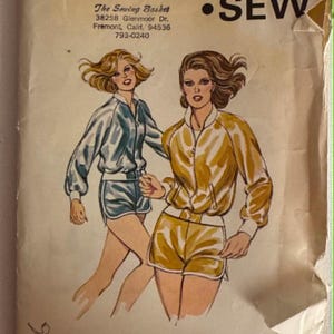 May include: A Kwik Sew pattern envelope, number 933, for sizes XS-S-M-L. The illustration shows two individuals wearing matching zip-up jackets and shorts sets, one in blue and one in yellow, with white trim. The paper envelope is worn and creased.