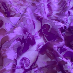 May include: Close-up of a flowing fabric with a floral pattern. The fabric is a vibrant purple with darker purple and light brown floral designs. The material appears soft and has a slight sheen, with visible folds and textures.
