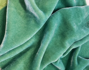 May include: A close-up of a soft, green velvet fabric with a subtle sheen. The fabric is folded and draped, showcasing its texture and drape.