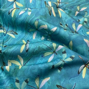 May include: Close-up of a teal fabric with a repeating dragonfly pattern. The dragonflies have yellow, white, and black details. The fabric has a marbled effect, creating a flowing, textured appearance. Ideal for sewing projects.