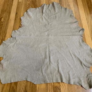 May include: A large, irregularly shaped piece of light tan leather with a textured surface. The edges are uneven, and the leather appears to be a single hide. The material is lying on a wooden floor.
