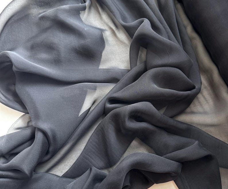 May include: Black sheer fabric with a soft, flowing texture. The fabric is draped and folded, showcasing its delicate and translucent quality.