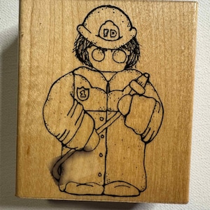 Puede incluir: Un sello de madera con un bombero de dibujos animados sosteniendo una manguera. El bombero lleva un casco con las letras "FD" y un uniforme con el número "1". El sello es de madera clara.