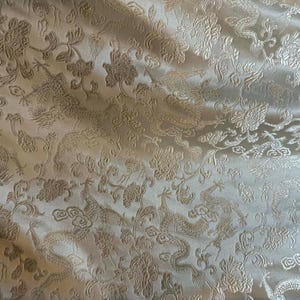 Puede incluir: Tela de color crema con un estampado en relieve de dragones y flores. El diseño presenta dragones, flores y motivos de nubes arremolinadas. La tela tiene un brillo sutil, creando una apariencia lujosa. Este textil es adecuado para varios proyectos de artesanía.