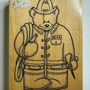 Puede incluir: Un sello de madera con un oso vestido de bombero. El oso lleva un casco con la inscripción "FIRE DEPT" y un uniforme con la palabra "BEAR" en un bolsillo. El oso sostiene una manguera y un hacha. El sello está sobre un bloque de madera marrón claro.