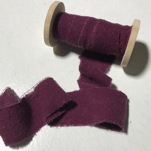 May include: A spool of deep burgundy fabric ribbon with a raw edge. The ribbon is wound around a wooden spool.