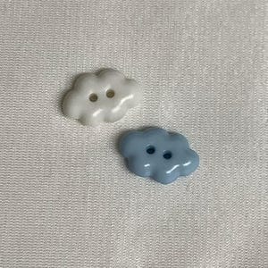 以下が含まれることがあります： 雲の形をしたボタンが2つ。1つは白、もう1つは水色です。各ボタンには縫い付け用の穴が2つあります。ボタンは光沢のある素材でできており、白いテクスチャの背景に配置されています。