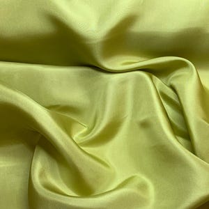 Puede incluir: Una tela de color verde lima sólido con un acabado suave y lustroso. El material está drapeado en pliegues suaves y fluidos que crean sombras profundas y reflejos brillantes en la superficie, enfatizando la calidad reflectante del textil.