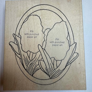 May include: A wooden stamp featuring a floral design with two tulip-like flowers inside an oval frame. The text "Fill with punched paper art" is printed on the stamp. The stamp is made of light-colored wood.