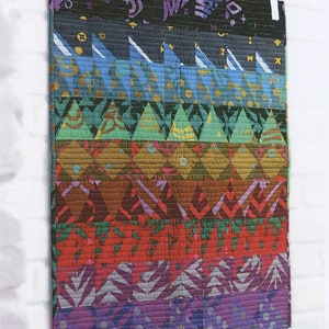 May include: A colorful quilt pattern called "Gonzo" by Needle in a Hayes Stack. The quilt is made up of a series of colorful triangles and squares in a variety of colors, including red, orange, yellow, green, blue, purple, and brown. The quilt is hanging on a white brick wall. The pattern number is NH1616.