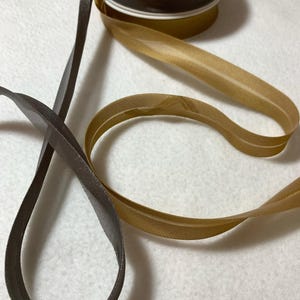 Könnte beinhalten: Zwei Rollen Band, eine grau und eine goldfarben, werden auf einer weißen Oberfläche gezeigt. Das graue Band ist einfarbig, während das goldene Band einen dezenten Glanz und ein strukturiertes Muster hat.