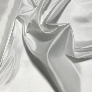 Puede incluir: Tela de satén blanco con una textura suave y sedosa. La tela está drapeada y doblada, creando curvas y pliegues suaves.