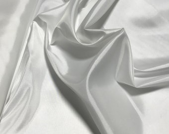 May include: White satin fabric with a smooth, silky texture. The fabric is draped and folded, creating soft curves and folds.