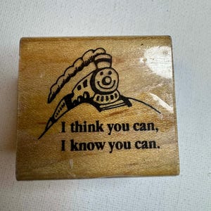 May include: Wooden stamp featuring a smiling cartoon train with the text "I think you can, I know you can." The stamp is rectangular and made of light-colored wood, with the train design and text in black.