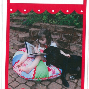 May include: A young girl sits on a colorful patchwork pillow reading a book with a black dog beside her. The pillow is labeled "Perfect Pillow Poof" and the image is from "Little Lambs".
