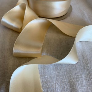 Könnte beinhalten: Eine Spule cremefarbenes Seidensatinband mit glänzendem Glanz. Das Band ist teilweise abgerollt und zeigt seine glatte Textur und den eleganten Fall. Das Etikett der Spule ist sichtbar und gibt die Marke und Art des Bandes an.