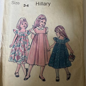 May include: Vintage sewing pattern titled "The Children's Corner Patterns 98 Hillary" for sizes 3-4. The illustration shows three girls in dresses, with views A, B, and C labeled below. The dresses are in pink, blue, and floral patterns.