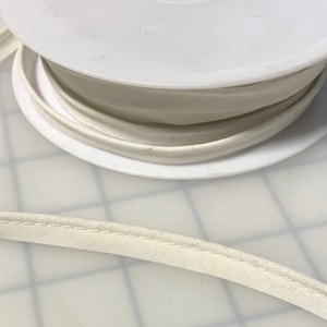 May include: A spool of white bias tape with a white stitching detail. The tape is wound around a white plastic spool.