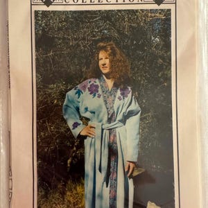 May include: A sewing pattern envelope titled The Patchwork Collection Pansy Robe. The cover features a light blue robe with purple pansy floral appliques on the shoulders and a patterned trim along the front opening, tied with a matching belt.