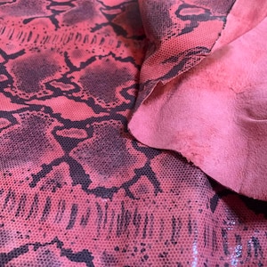 Pode incluir: Close-up de um pedaço de couro de pele de cobra rosa com um padrão escuro. O couro é dobrado, revelando uma parte inferior de camurça rosa macia. O padrão de pele de cobra é um design repetitivo de formas escuras.