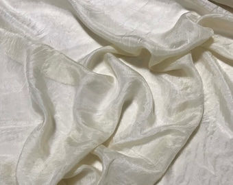 Op de afbeelding: Een close-up van een witte zijden stof met een gladde, vloeiende textuur. De stof is gedrapeerd en gevouwen, waardoor zachte, afgeronde vormen ontstaan.
