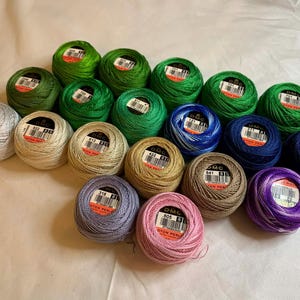 May include: A collection of DMC Cotton Perle thread balls in various colors, including green, blue, pink, and gold. Each ball has a label with a product code. The threads are arranged on a white surface.
