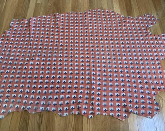 May include: A large, irregularly shaped piece of fabric with a repeating pattern of white crosses within blue squares on a coral background. The fabric is lying on a wooden surface.