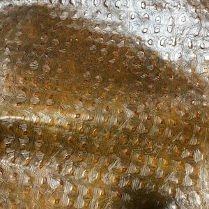 May include: A close-up of a textured gold fabric with a repeating pattern of small hexagons. The fabric has a subtle sheen and a slightly raised texture.